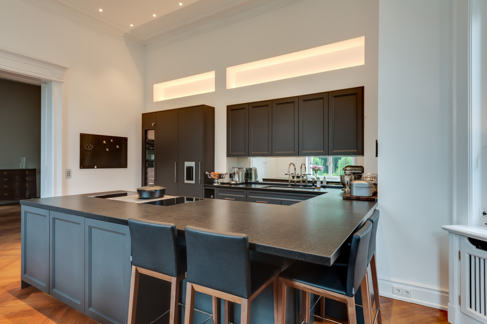 Photo of an expansive midcentury u-shaped open plan kitchen in Hamburg with a double-bowl sink, beaded inset cabinets, black cabinets, laminate benchtops, white splashback, mirror splashback, black appliances, painted wood floors, a peninsula, brown floor, black benchtop and vaulted.