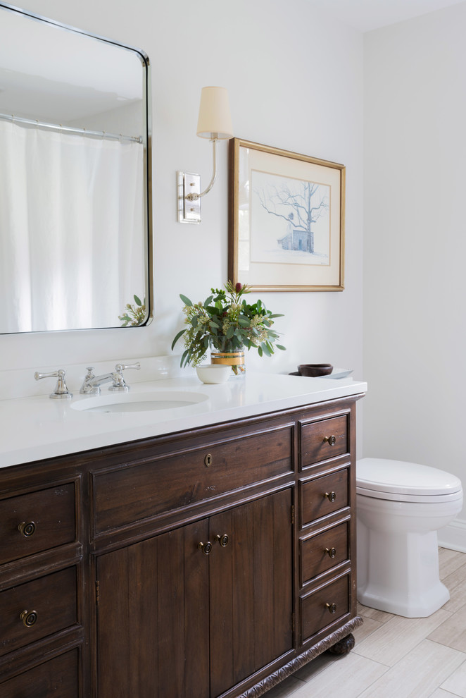 This is an example of a mid-sized transitional bathroom in Other with furniture-like cabinets, dark wood cabinets, a drop-in tub, a one-piece toilet, white tile, subway tile, white walls, limestone floors, a console sink, engineered quartz benchtops and beige floor.
