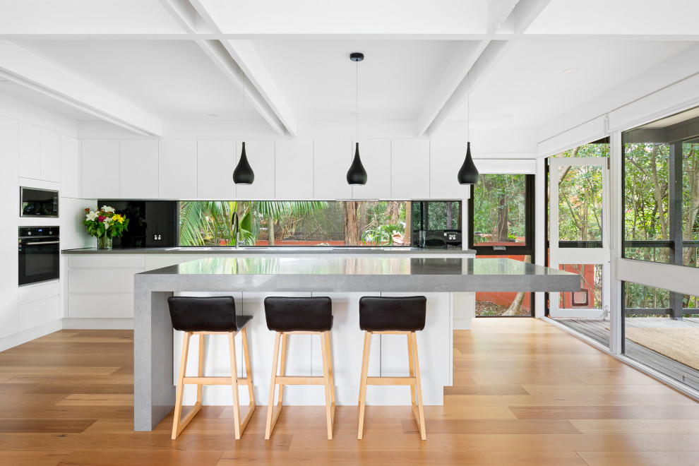 Photo of a mid-sized modern l-shaped eat-in kitchen in Sydney with an undermount sink, white cabinets, solid surface benchtops, black splashback, mirror splashback, black appliances, medium hardwood floors, with island, brown floor, grey benchtop and exposed beam.