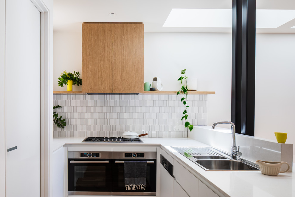 This is an example of a contemporary u-shaped kitchen in Melbourne with a drop-in sink, flat-panel cabinets, white cabinets, white splashback, mosaic tile splashback, stainless steel appliances and white benchtop.