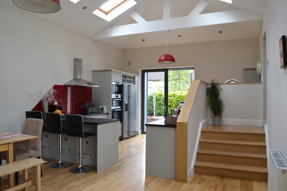 This is an example of a mid-sized modern l-shaped open plan kitchen in Dublin with a drop-in sink, flat-panel cabinets, grey cabinets, laminate benchtops, red splashback, glass sheet splashback, stainless steel appliances, light hardwood floors and no island.