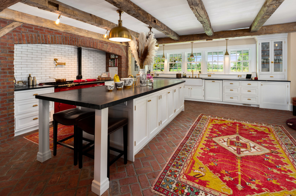 Réalisation d'une cuisine champêtre en L avec un évier de ferme, un placard avec porte à panneau encastré, des portes de placard blanches, un électroménager de couleur, un sol en brique, îlot, un sol rouge, plan de travail noir et poutres apparentes.