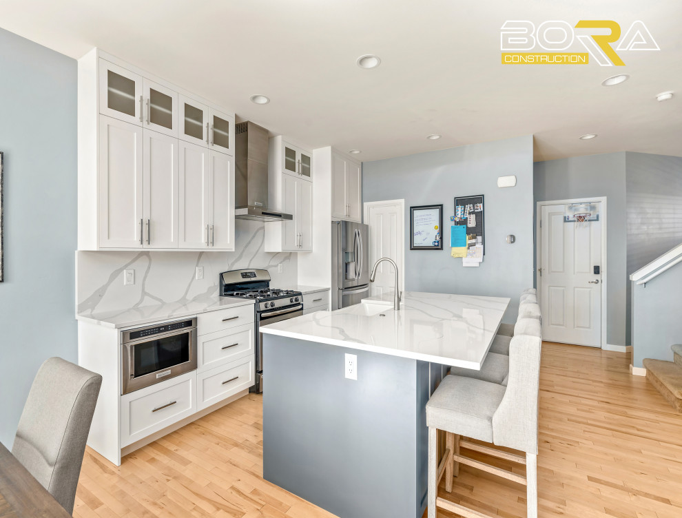 Example of a mid-sized single-wall light wood floor and brown floor eat-in kitchen design in Seattle with a farmhouse sink, shaker cabinets, white cabinets, quartz countertops, white backsplash, quartz backsplash, stainless steel appliances, an island and white countertops