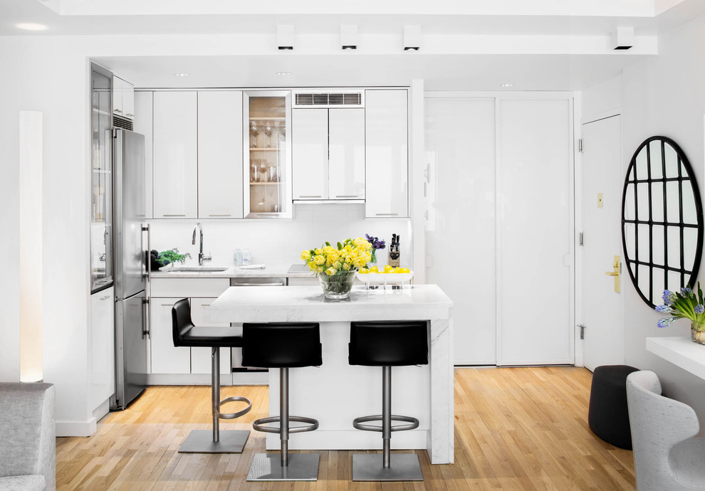 Small contemporary l-shaped open plan kitchen in New York with an undermount sink, flat-panel cabinets, white cabinets, marble benchtops, white splashback, stainless steel appliances, light hardwood floors, with island and brown floor.