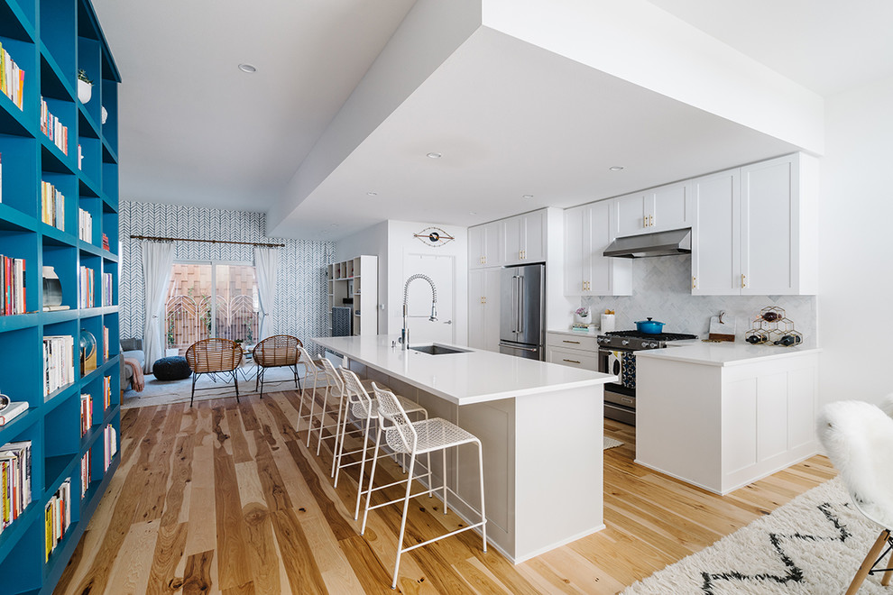 Large scandinavian galley open plan kitchen in Austin with light hardwood floors, an undermount sink, shaker cabinets, white cabinets, solid surface benchtops, white splashback, porcelain splashback, stainless steel appliances and with island.