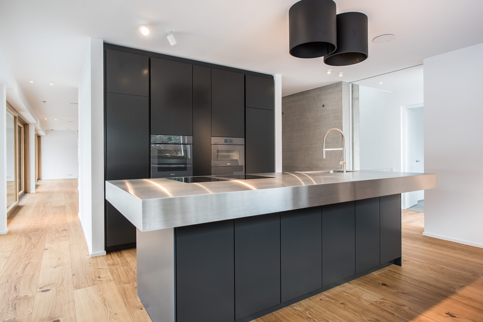 Photo of a mid-sized contemporary galley open plan kitchen in Stuttgart with an integrated sink, flat-panel cabinets, black cabinets, stainless steel benchtops, stainless steel appliances, medium hardwood floors, with island, brown floor and grey benchtop.
