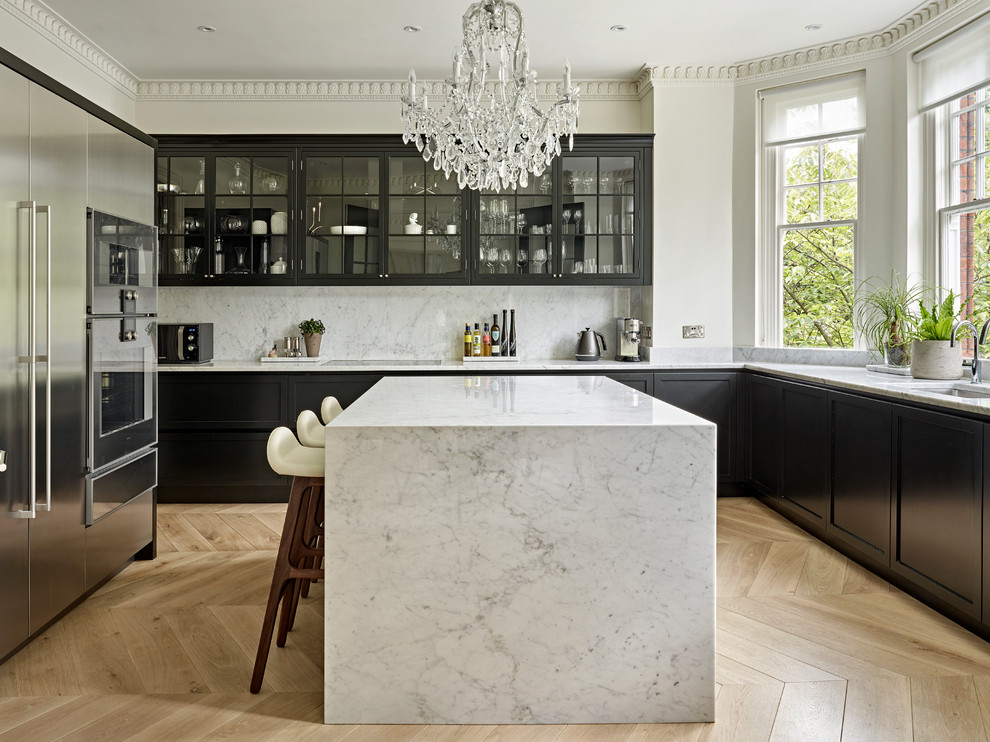 This is an example of a transitional u-shaped kitchen in London with an undermount sink, shaker cabinets, black cabinets, white splashback, stone slab splashback, stainless steel appliances, light hardwood floors, with island, beige floor and white benchtop.