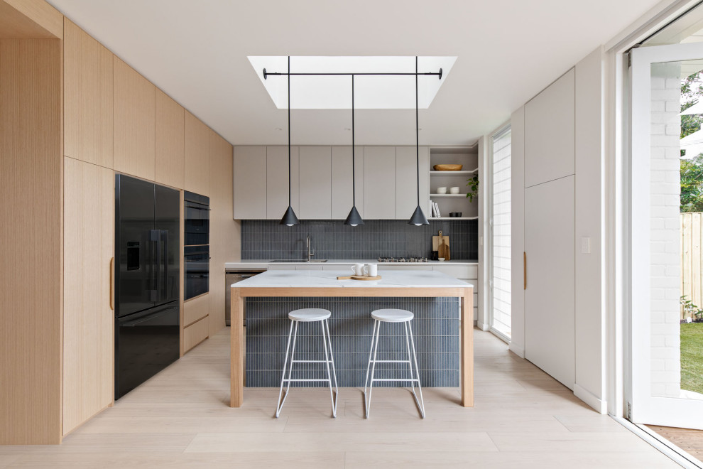 This is an example of a large modern l-shaped eat-in kitchen in Sydney with light wood cabinets, solid surface benchtops, blue splashback, ceramic splashback, black appliances, light hardwood floors, with island, white benchtop, flat-panel cabinets and beige floor.