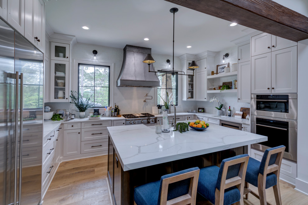 This is an example of a transitional u-shaped kitchen in Denver with shaker cabinets, white cabinets, white splashback, stainless steel appliances, light hardwood floors, with island and white benchtop.
