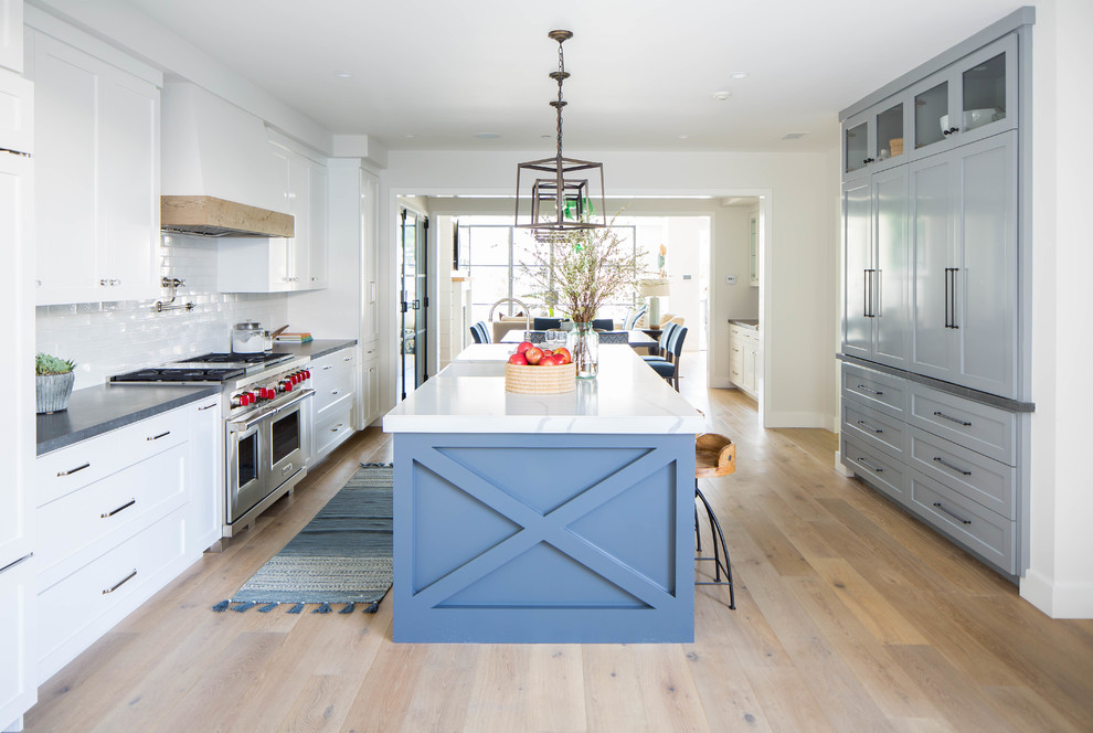 This is an example of a large country galley open plan kitchen in Orange County with a farmhouse sink, shaker cabinets, white cabinets, white splashback, subway tile splashback, stainless steel appliances, medium hardwood floors, with island, brown floor and white benchtop.
