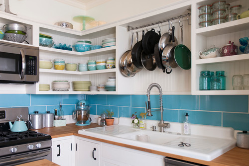 Bright kitchen with open kitchen cabinet shelves displaying colorful dishes and glass jars, teal tile backsplash, hanging pots, and a farmhouse sink.