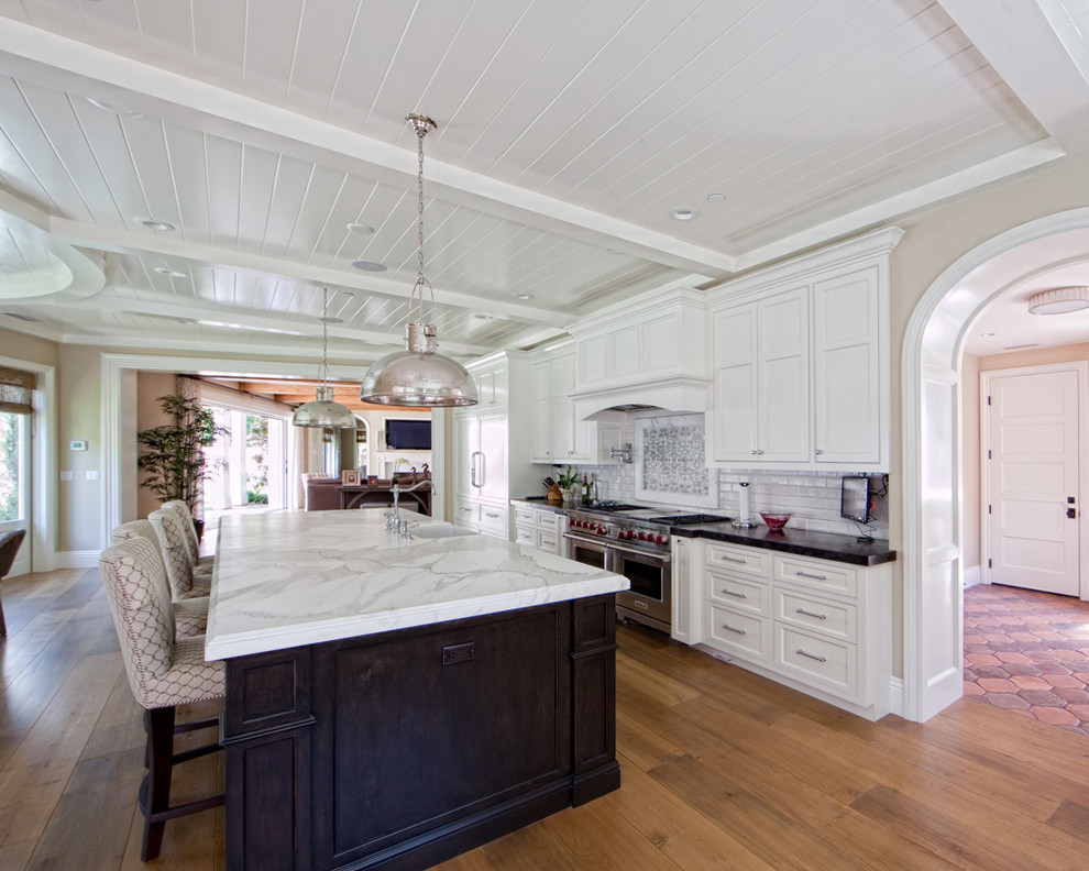 This is an example of a mid-sized traditional single-wall separate kitchen in Orange County with a farmhouse sink, shaker cabinets, white cabinets, marble benchtops, white splashback, subway tile splashback, stainless steel appliances, medium hardwood floors, with island and brown floor.