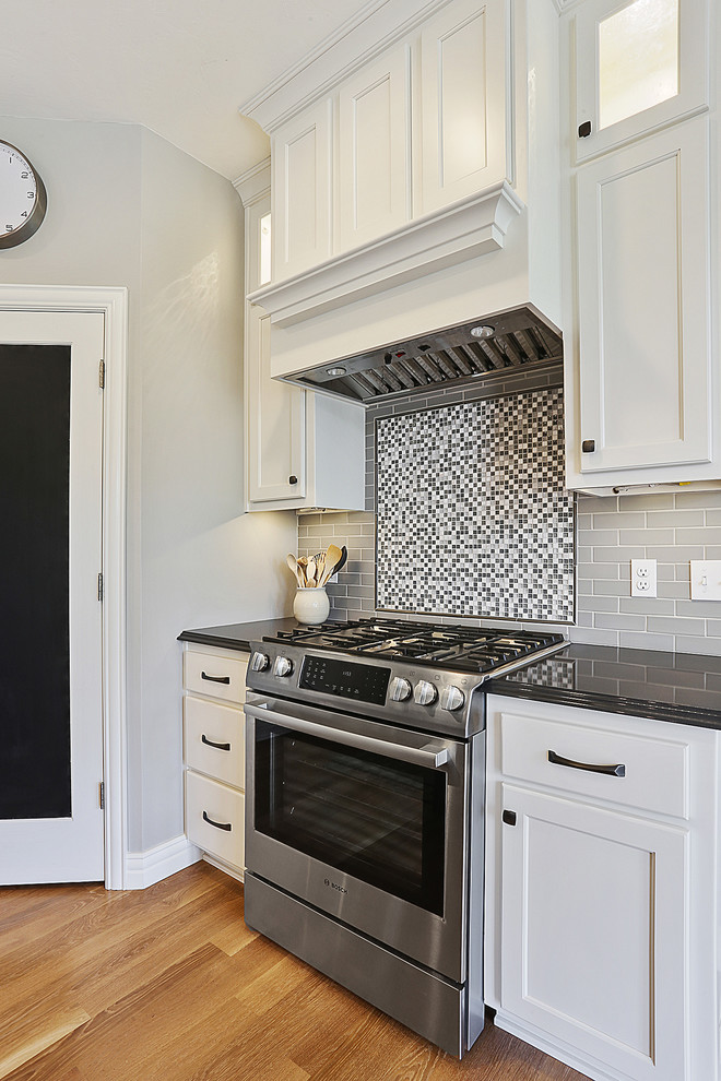 Example of a galley medium tone wood floor and brown floor kitchen design with white cabinets, multicolored backsplash, stainless steel appliances, an island, an undermount sink and mosaic tile backsplash