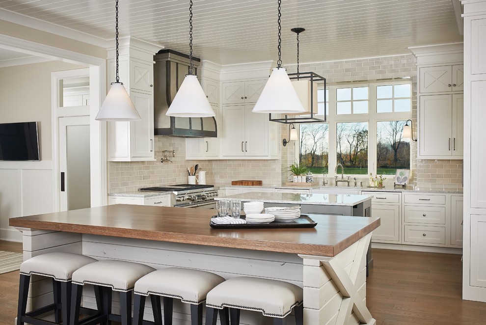 This is an example of a beach style u-shaped kitchen with white cabinets, beige splashback, medium hardwood floors, multiple islands, brown floor, a farmhouse sink, shaker cabinets, wood benchtops, subway tile splashback and brown benchtop.