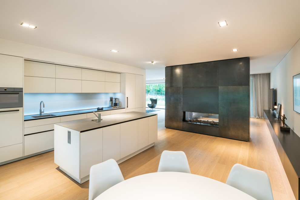 This is an example of a modern galley open plan kitchen in Frankfurt with an undermount sink, flat-panel cabinets, white cabinets, stainless steel appliances, light hardwood floors, with island, beige floor and grey benchtop.