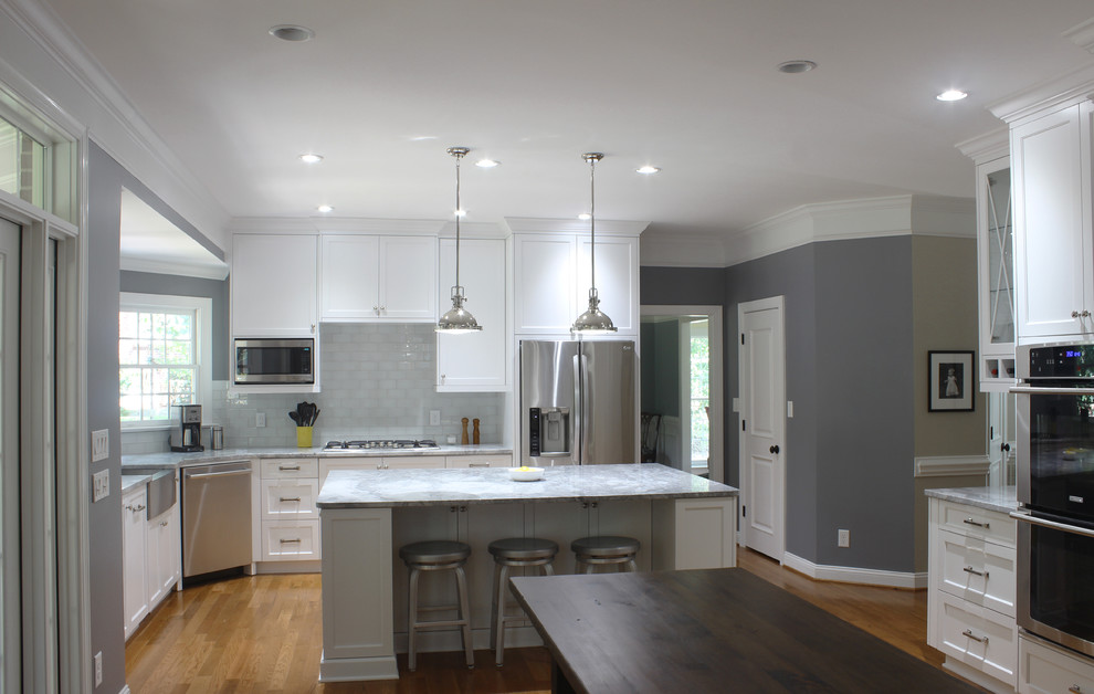Photo of a mid-sized transitional l-shaped eat-in kitchen in Charlotte with a farmhouse sink, recessed-panel cabinets, white cabinets, marble benchtops, grey splashback, subway tile splashback, stainless steel appliances, medium hardwood floors and with island.
