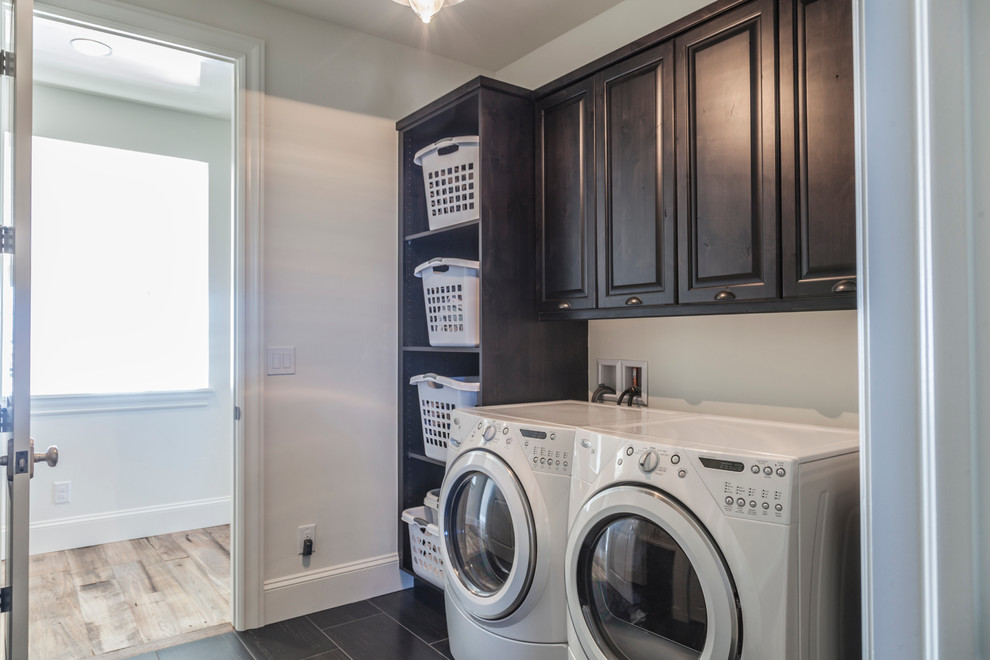 Photo of a mid-sized industrial single-wall dedicated laundry room in Other with raised-panel cabinets, grey walls, porcelain floors, a side-by-side washer and dryer and dark wood cabinets.