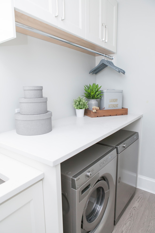 This is an example of a mid-sized beach style single-wall dedicated laundry room in Vancouver with an undermount sink, shaker cabinets, white cabinets, grey walls, light hardwood floors, a side-by-side washer and dryer and grey floor.