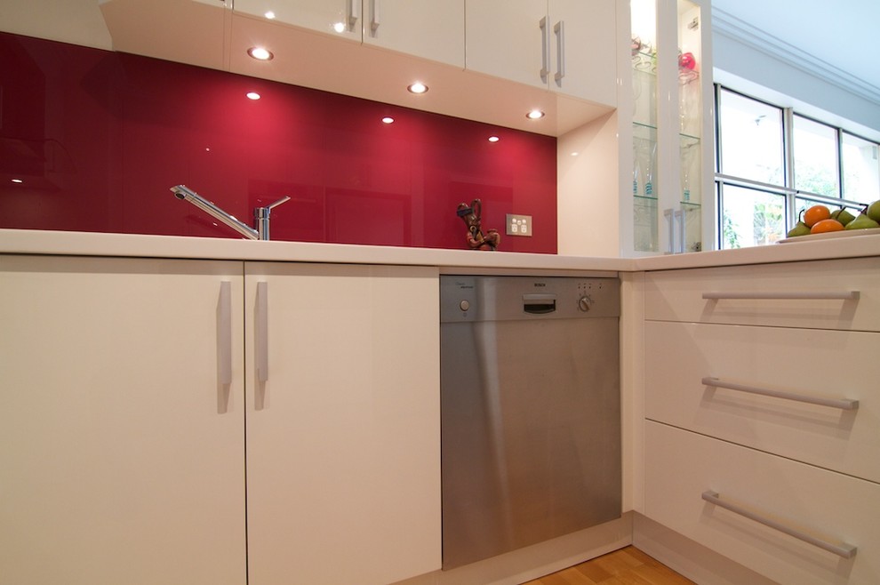 Design ideas for a small modern u-shaped separate kitchen in Sydney with an integrated sink, flat-panel cabinets, white cabinets, solid surface benchtops, red splashback, glass sheet splashback, stainless steel appliances, light hardwood floors and no island.