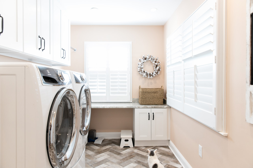 This is an example of a mid-sized modern u-shaped utility room in DC Metro with beaded inset cabinets, white cabinets, marble benchtops, pink walls, light hardwood floors, a stacked washer and dryer, multi-coloured floor and beige benchtop.