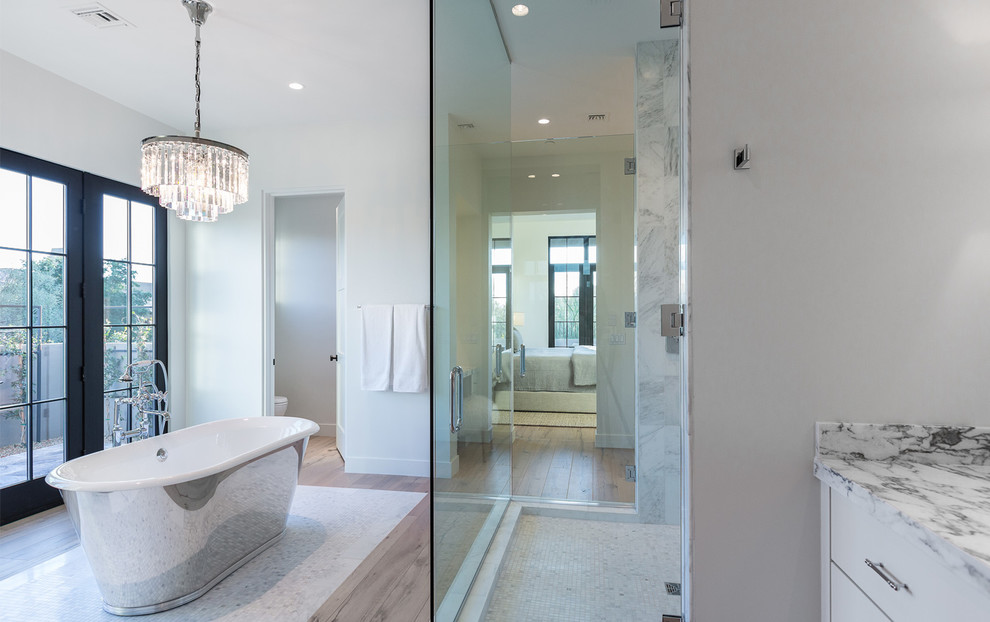 This is an example of a large transitional master bathroom in Tampa with flat-panel cabinets, white cabinets, a freestanding tub, an alcove shower, gray tile, white tile, white walls, light hardwood floors, a hinged shower door, marble, marble benchtops and brown floor.