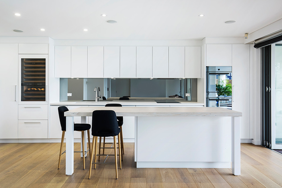 This is an example of a contemporary galley kitchen in Sydney with an undermount sink, flat-panel cabinets, white cabinets, glass sheet splashback, stainless steel appliances, light hardwood floors, with island and beige floor.