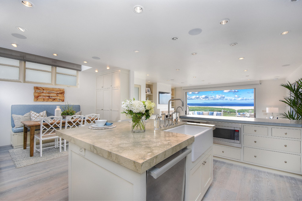 Mid-sized beach style u-shaped open plan kitchen in Orange County with a farmhouse sink, glass-front cabinets, white cabinets, grey splashback, matchstick tile splashback, stainless steel appliances, with island, grey floor and light hardwood floors.