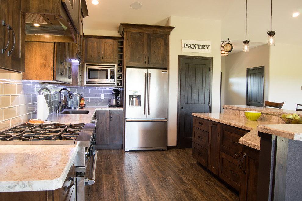 Example of a mountain style l-shaped brown floor eat-in kitchen design with shaker cabinets, brown cabinets, laminate countertops, gray backsplash, stainless steel appliances, an island and beige countertops