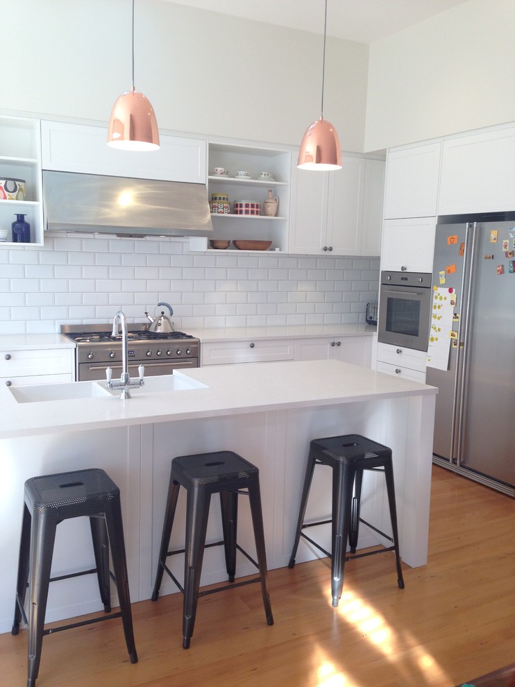 Photo of a large transitional l-shaped open plan kitchen in Auckland with a farmhouse sink, recessed-panel cabinets, white cabinets, quartz benchtops, white splashback, subway tile splashback, stainless steel appliances, medium hardwood floors and with island.