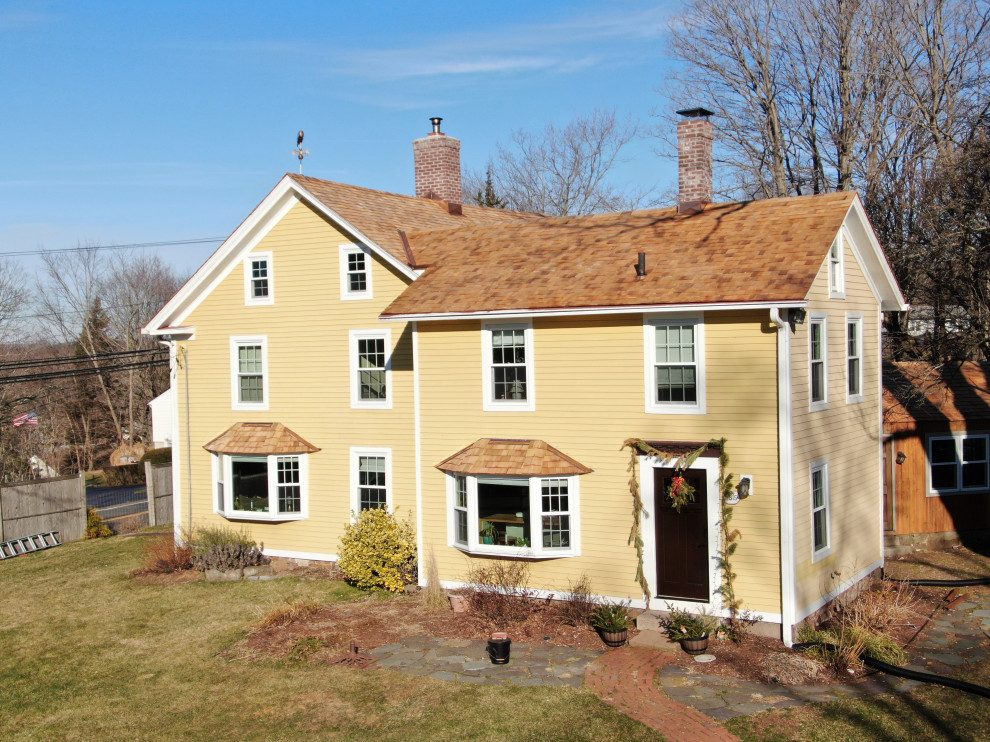 Foto della villa ampia gialla country a tre piani con rivestimento in legno, tetto a capanna, copertura a scandole, tetto rosso e pannelli sovrapposti