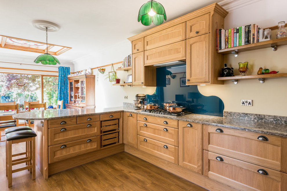 This is an example of a traditional l-shaped eat-in kitchen in Other with recessed-panel cabinets, medium wood cabinets, blue splashback, glass sheet splashback, medium hardwood floors and multi-coloured benchtop.