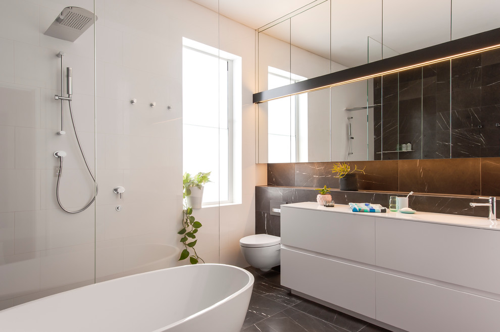 Photo of a mid-sized contemporary 3/4 bathroom in Sydney with furniture-like cabinets, white cabinets, a freestanding tub, an open shower, a wall-mount toilet, gray tile, stone tile, white walls, marble floors, an integrated sink and solid surface benchtops.