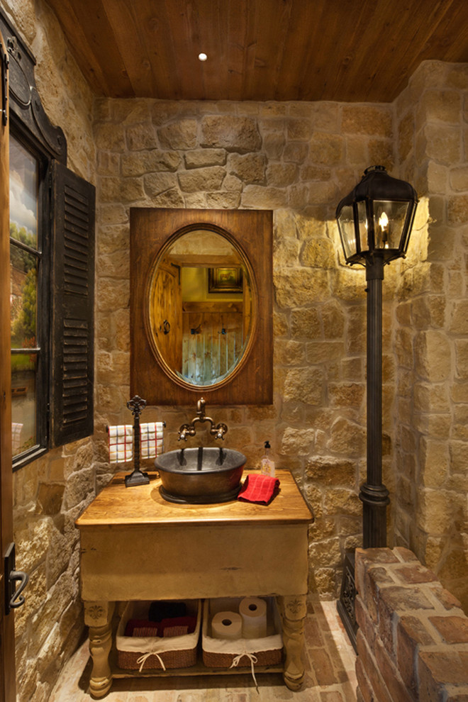 Mid-sized mediterranean powder room in Dallas with open cabinets, a vessel sink, wood benchtops, light wood cabinets, a two-piece toilet, beige tile, stone tile, beige walls, brick floors, beige floor and brown benchtops.