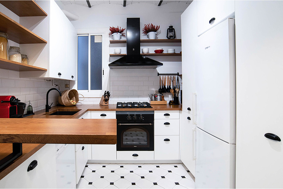 Small country u-shaped open plan kitchen in Barcelona with a single-bowl sink, flat-panel cabinets, white cabinets, wood benchtops, black splashback, black appliances, ceramic floors, no island, white floor, brown benchtop and vaulted.