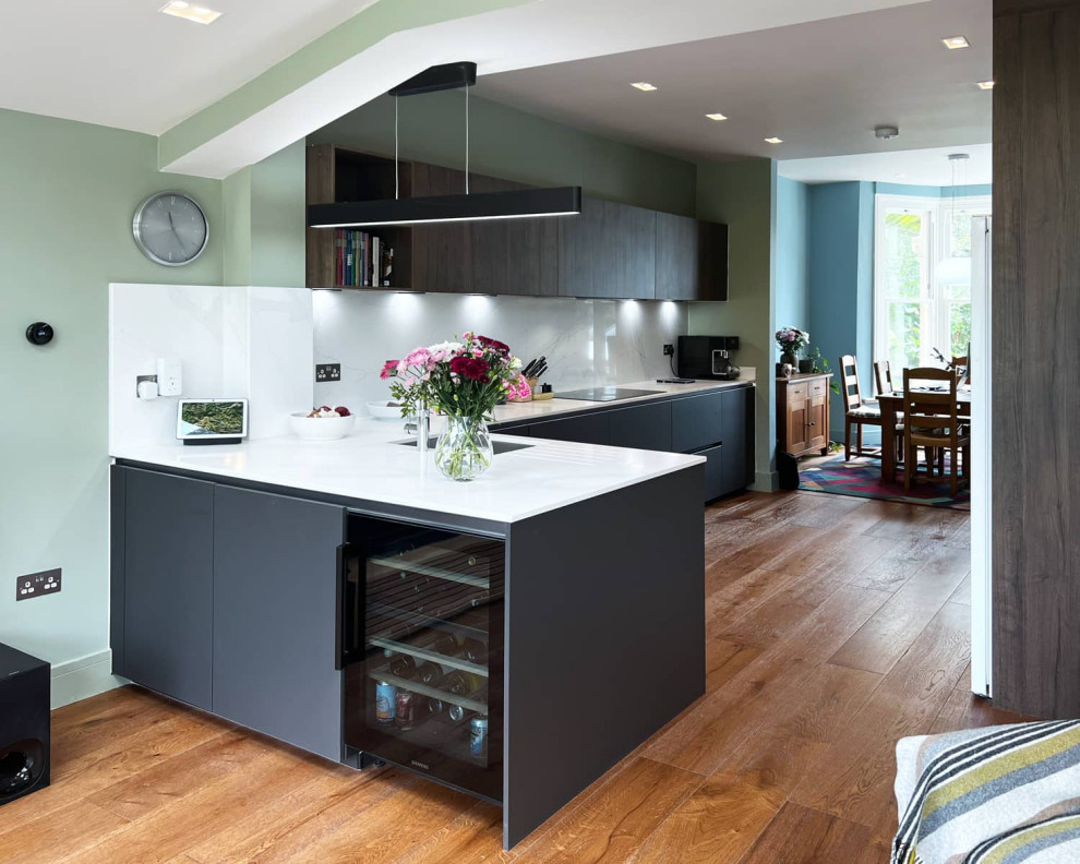 This is an example of a mid-sized contemporary galley eat-in kitchen in London with a single-bowl sink, flat-panel cabinets, grey cabinets, quartzite benchtops, white splashback, black appliances, light hardwood floors, brown floor and white benchtop.