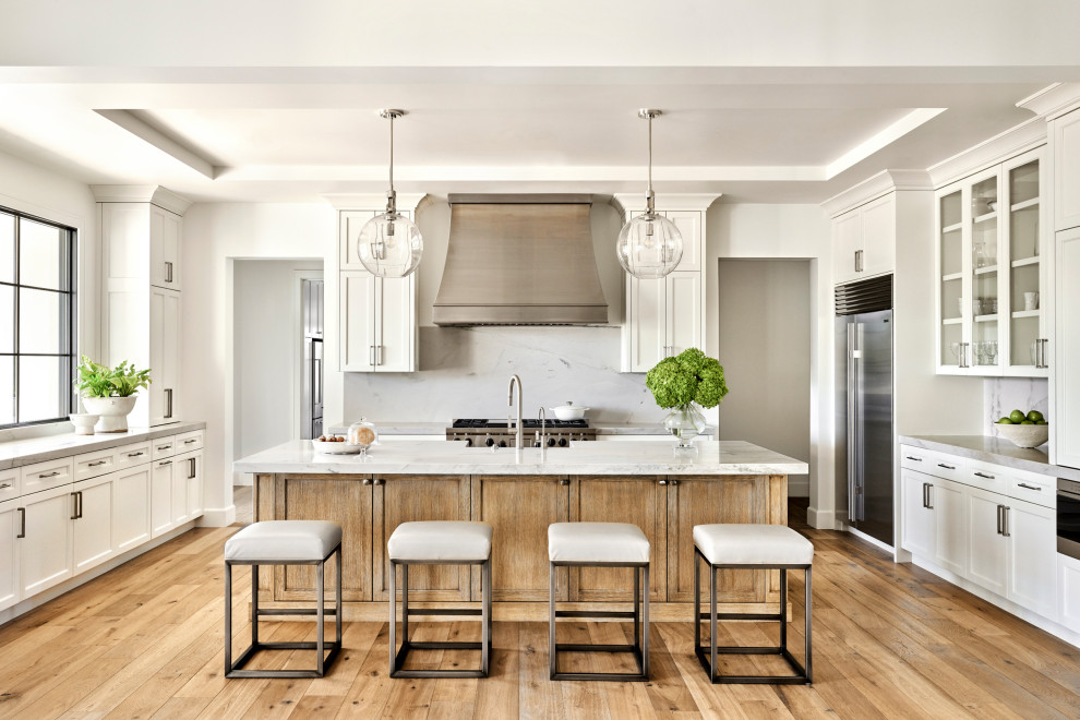 Large transitional u-shaped kitchen in Phoenix with a farmhouse sink, shaker cabinets, white cabinets, white splashback, stainless steel appliances, medium hardwood floors, with island, brown floor and white benchtop.