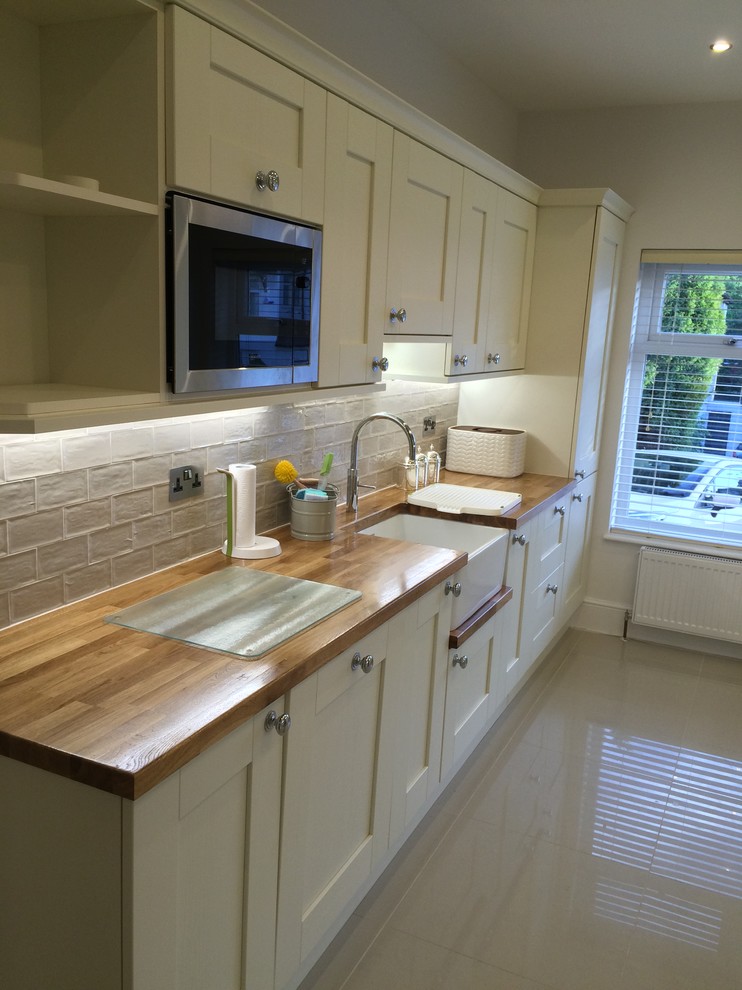 This is an example of a mid-sized country galley kitchen in Manchester with a farmhouse sink, shaker cabinets, wood benchtops, beige splashback, ceramic splashback, stainless steel appliances, porcelain floors and no island.