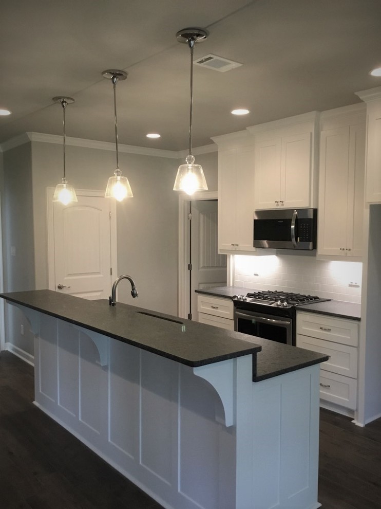This is an example of a small modern single-wall open plan kitchen in Little Rock with an undermount sink, shaker cabinets, white cabinets, granite benchtops, white splashback, ceramic splashback, stainless steel appliances, porcelain floors, with island, brown floor and black benchtop.
