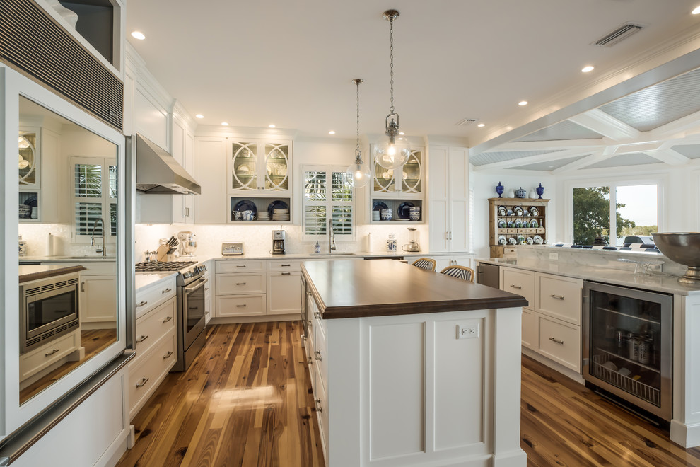 Photo of a beach style u-shaped kitchen in Miami with recessed-panel cabinets, white cabinets, white splashback, stainless steel appliances, dark hardwood floors, with island, brown floor and white benchtop.