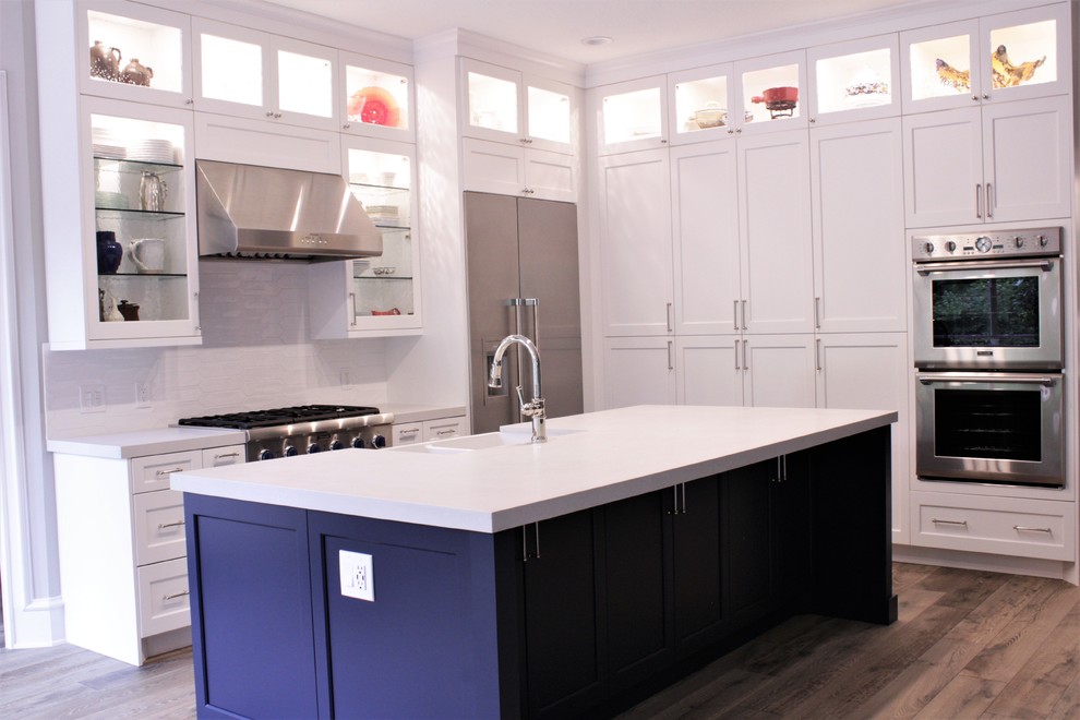 Photo of a mid-sized transitional l-shaped eat-in kitchen in Houston with a farmhouse sink, recessed-panel cabinets, white cabinets, quartz benchtops, white splashback, ceramic splashback, stainless steel appliances, light hardwood floors, with island, grey floor and white benchtop.