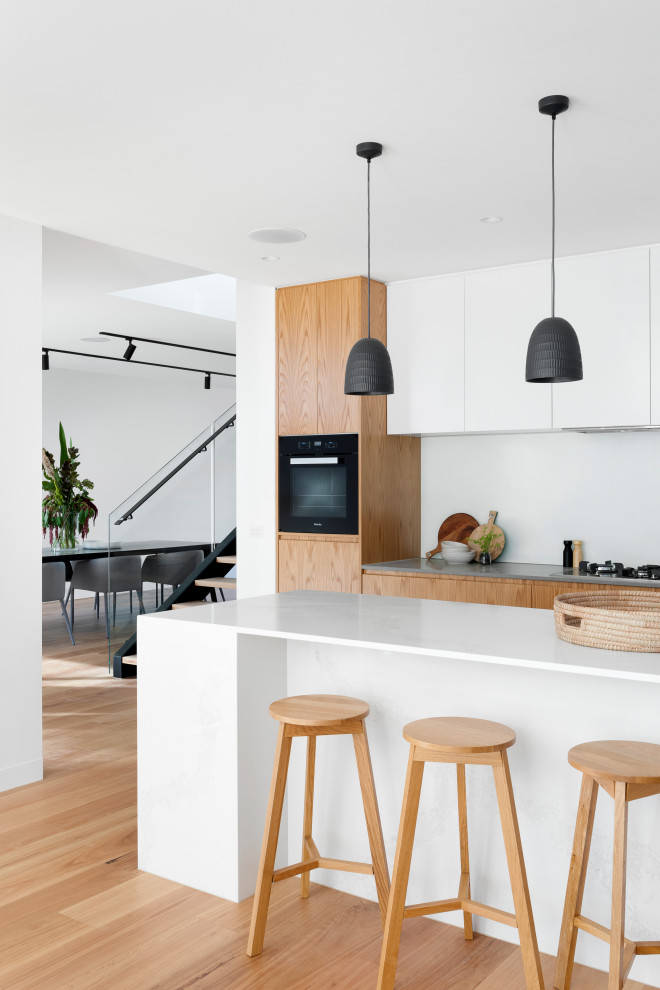 Design ideas for a mid-sized contemporary l-shaped open plan kitchen in Melbourne with an undermount sink, flat-panel cabinets, medium wood cabinets, quartz benchtops, white splashback, black appliances, medium hardwood floors, with island, brown floor and white benchtop.