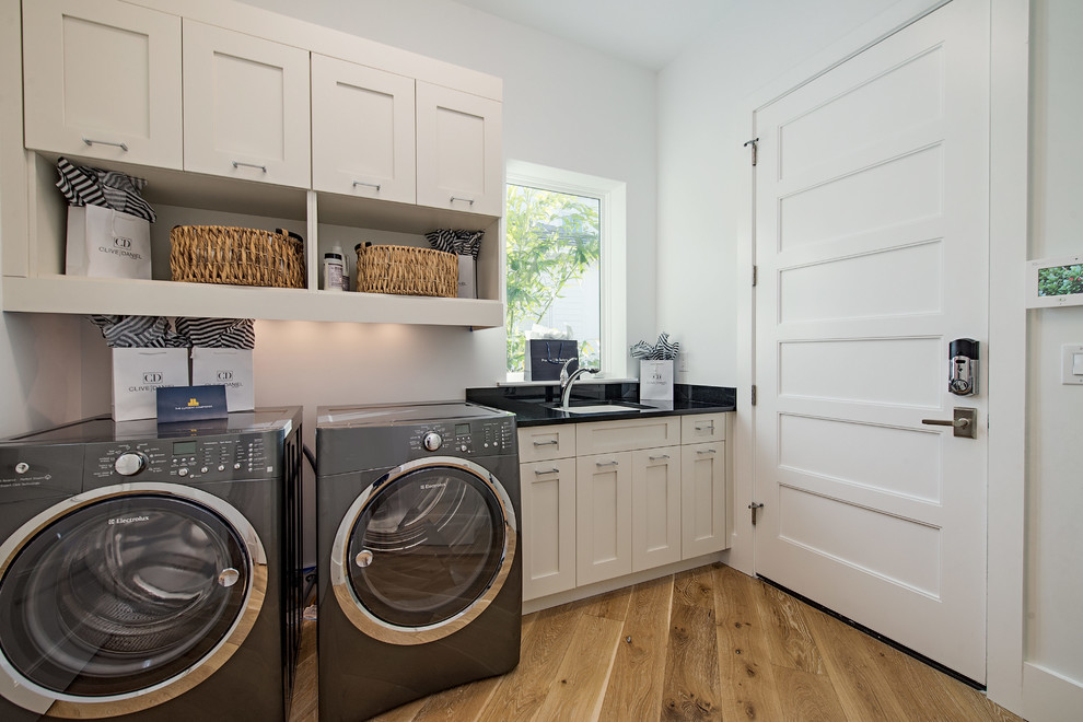 Inspiration for a transitional single-wall laundry room in Miami with an undermount sink, shaker cabinets, white cabinets, white walls, light hardwood floors, a side-by-side washer and dryer and black benchtop.