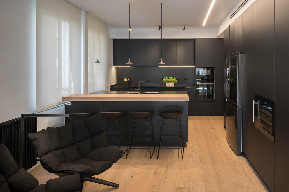 This is an example of a mid-sized contemporary l-shaped open plan kitchen in Barcelona with black cabinets, an undermount sink, flat-panel cabinets, black splashback, black appliances, light hardwood floors, a peninsula, beige floor and black benchtop.