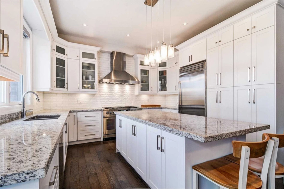 This is an example of a medium sized classic u-shaped kitchen/diner in Toronto with a submerged sink, shaker cabinets, white cabinets, granite worktops, white splashback, porcelain splashback, stainless steel appliances, dark hardwood flooring, an island, brown floors and white worktops.