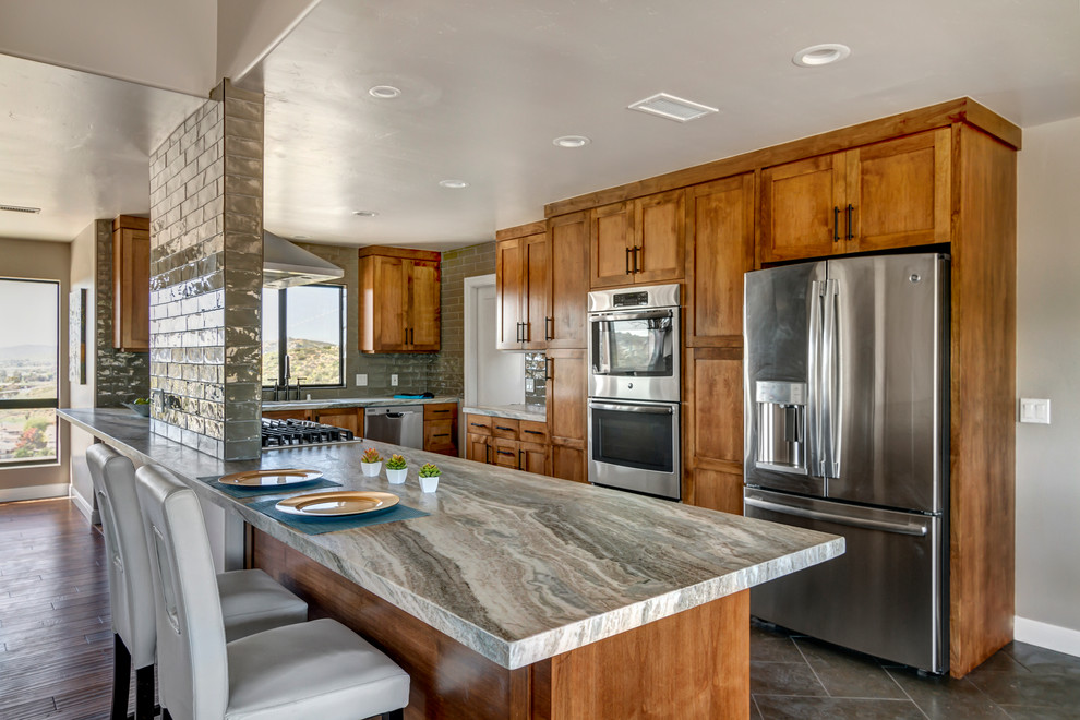 Photo of a large transitional l-shaped eat-in kitchen in San Diego with an undermount sink, shaker cabinets, brown cabinets, granite benchtops, grey splashback, ceramic splashback, stainless steel appliances, limestone floors and a peninsula.