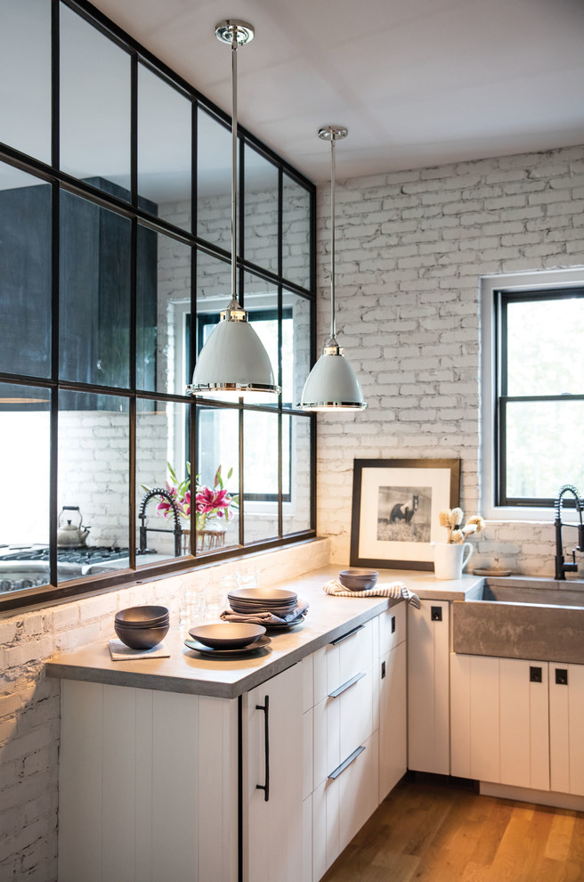 Photo of a mid-sized industrial l-shaped eat-in kitchen in Chicago with a farmhouse sink, louvered cabinets, white cabinets, solid surface benchtops, white splashback, brick splashback, stainless steel appliances, medium hardwood floors, a peninsula and brown floor.
