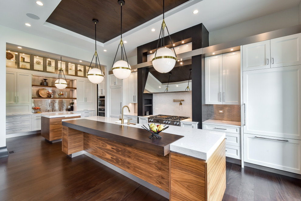 This is an example of a large modern kitchen in Chicago with a farmhouse sink, recessed-panel cabinets, white cabinets, tile benchtops, metallic splashback, metal splashback, white appliances, dark hardwood floors, multiple islands, brown floor, white benchtop and wood.