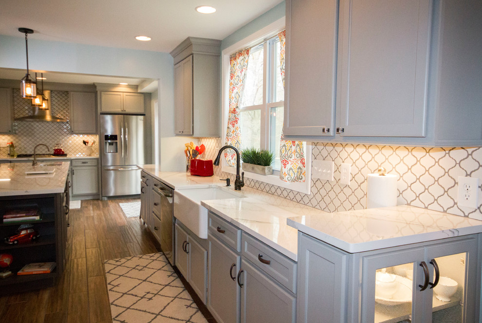 Example of a large transitional l-shaped medium tone wood floor and brown floor enclosed kitchen design in Cincinnati with a farmhouse sink, recessed-panel cabinets, gray cabinets, marble countertops, white backsplash, porcelain backsplash, stainless steel appliances and an island