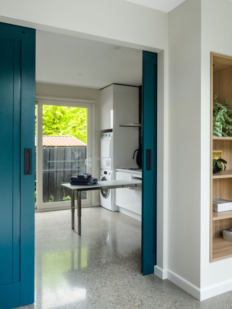 This is an example of a mid-sized contemporary laundry room in Melbourne with a single-bowl sink, open cabinets, medium wood cabinets, quartz benchtops, grey floor, grey benchtop, green splashback, ceramic splashback, white walls, concrete floors and a stacked washer and dryer.