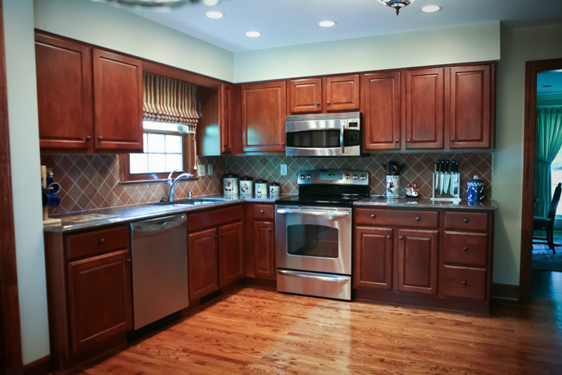 Design ideas for a mid-sized traditional l-shaped kitchen in Louisville with a double-bowl sink, raised-panel cabinets, medium wood cabinets, brown splashback, stainless steel appliances, medium hardwood floors, no island and brown floor.